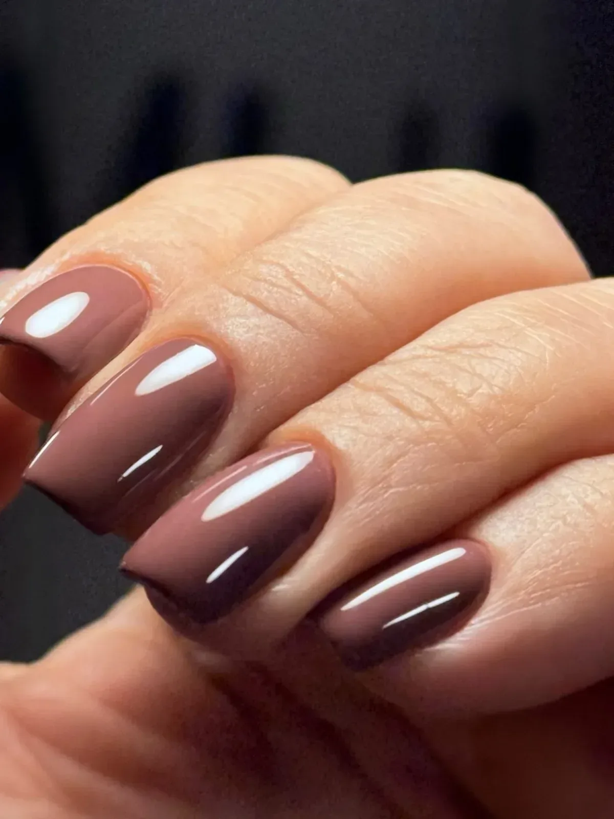Unhas esmaltadas em marrom acinzentado, fotografadas de perto sobre fundo escuro