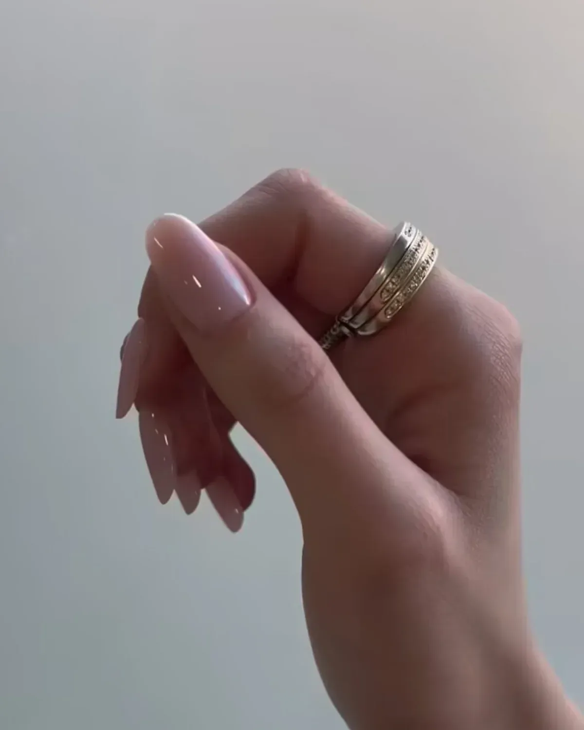 Mão com anéis e unhas nude recém-finalizadas, erguida contra um fundo claro